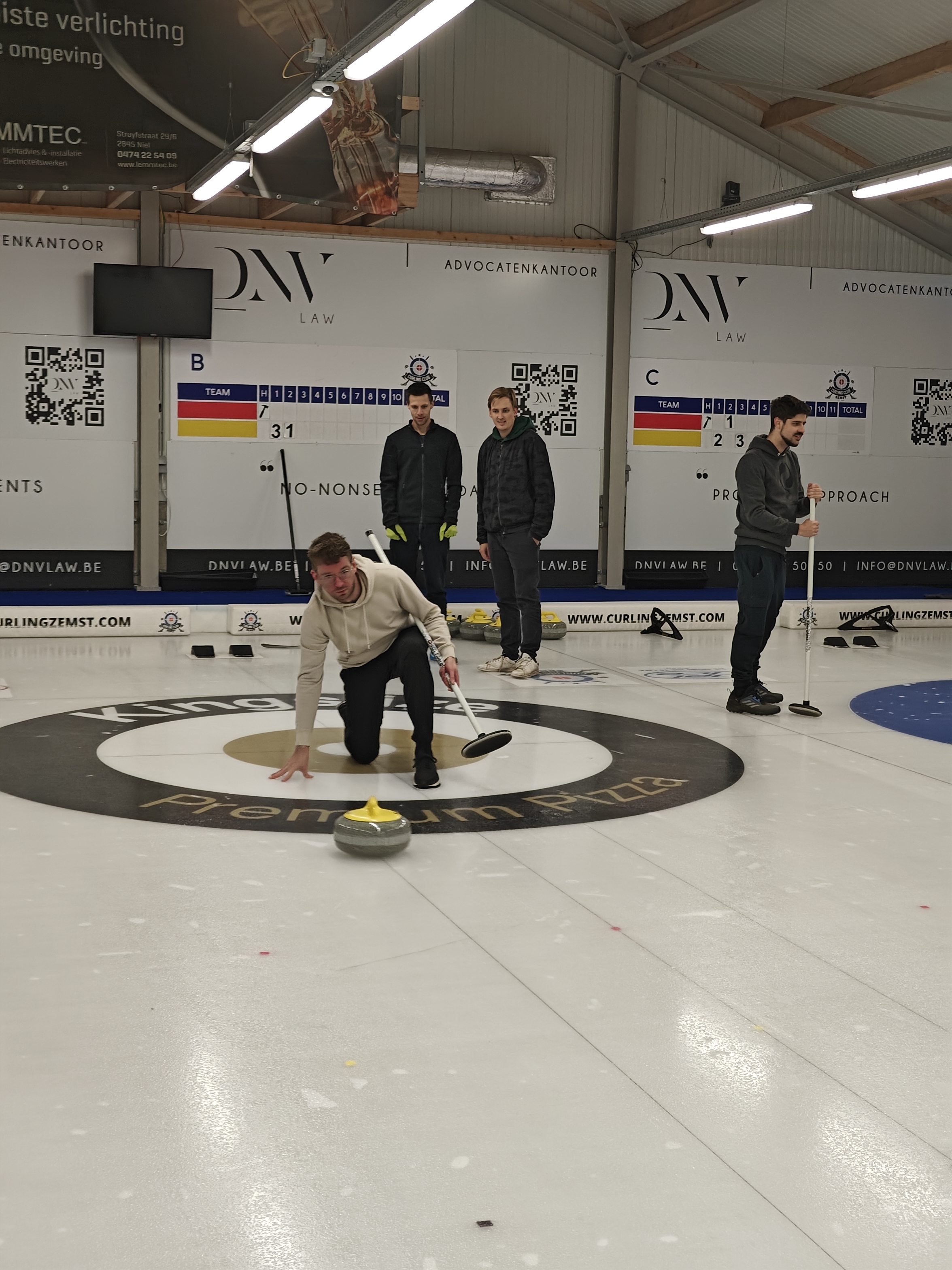 Itenium teammates bond over a competitive curling match, with one player expertly sliding a stone while others strategize, all set against a backdrop of scoreboards and local ads in a dedicated curling facility.