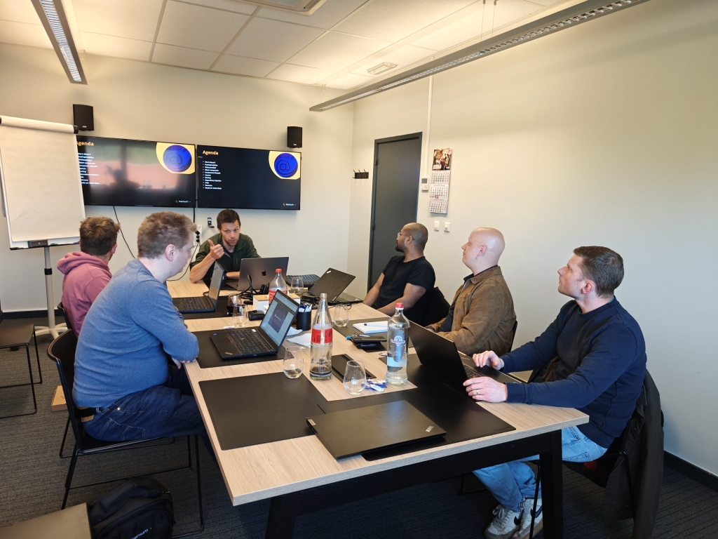 Attendees at a Blazor introduction session actively participating in a group discussion around a conference table, with laptops open and 'Agenda' displayed on dual monitors. 