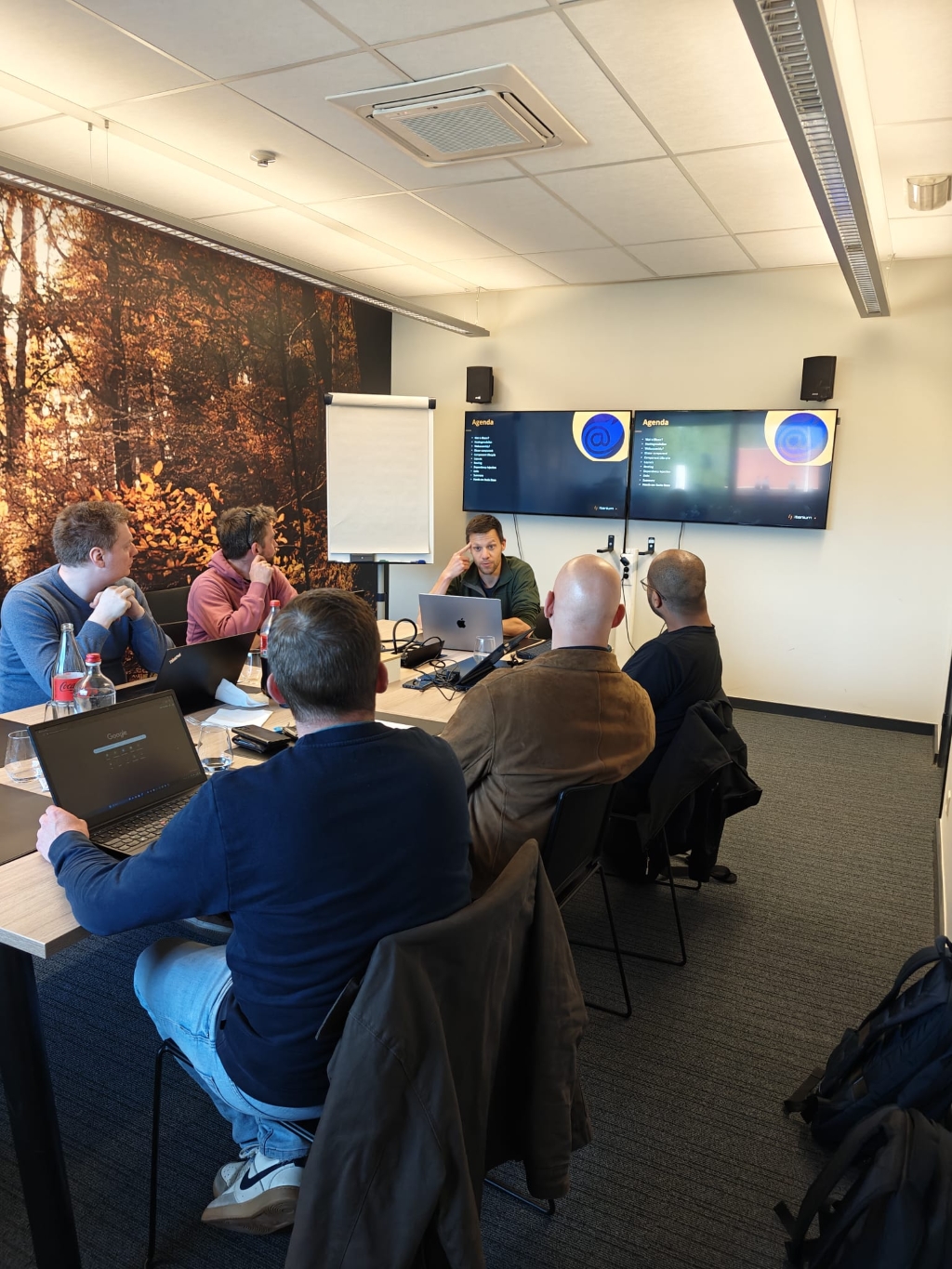 A group of software developers engrossed in a Blazor introductory session, seated around a conference table with laptops open and a presentation displayed on large screens. The room features a wall mural of a forest.