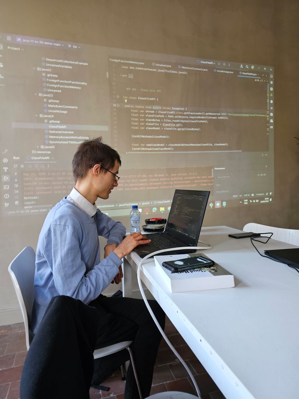 A focused developer, deeply engaged in a programming session, works on his laptop with Java code projected on the screen behind him, showcasing new features from Java 17 to 24 in a live demo. Perfect for a Java deep dive event.