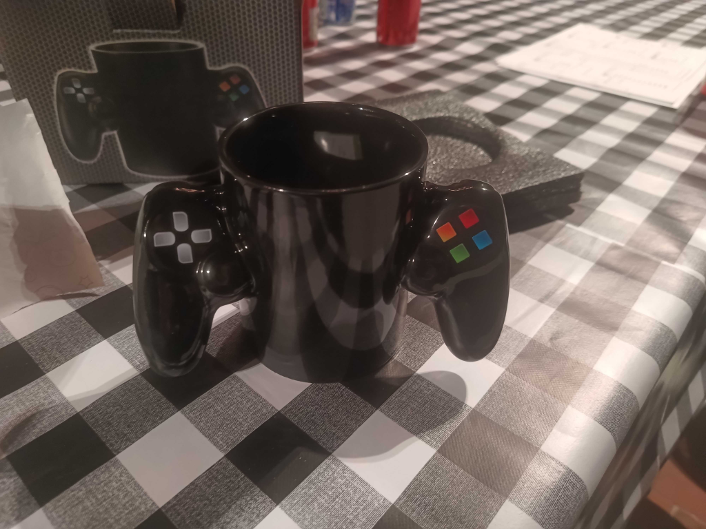 A black ceramic mug designed like a gaming controller, with handles mimicking a gamepad's D-pad and colorful buttons—a perfect prize for Michael's gaming victory.