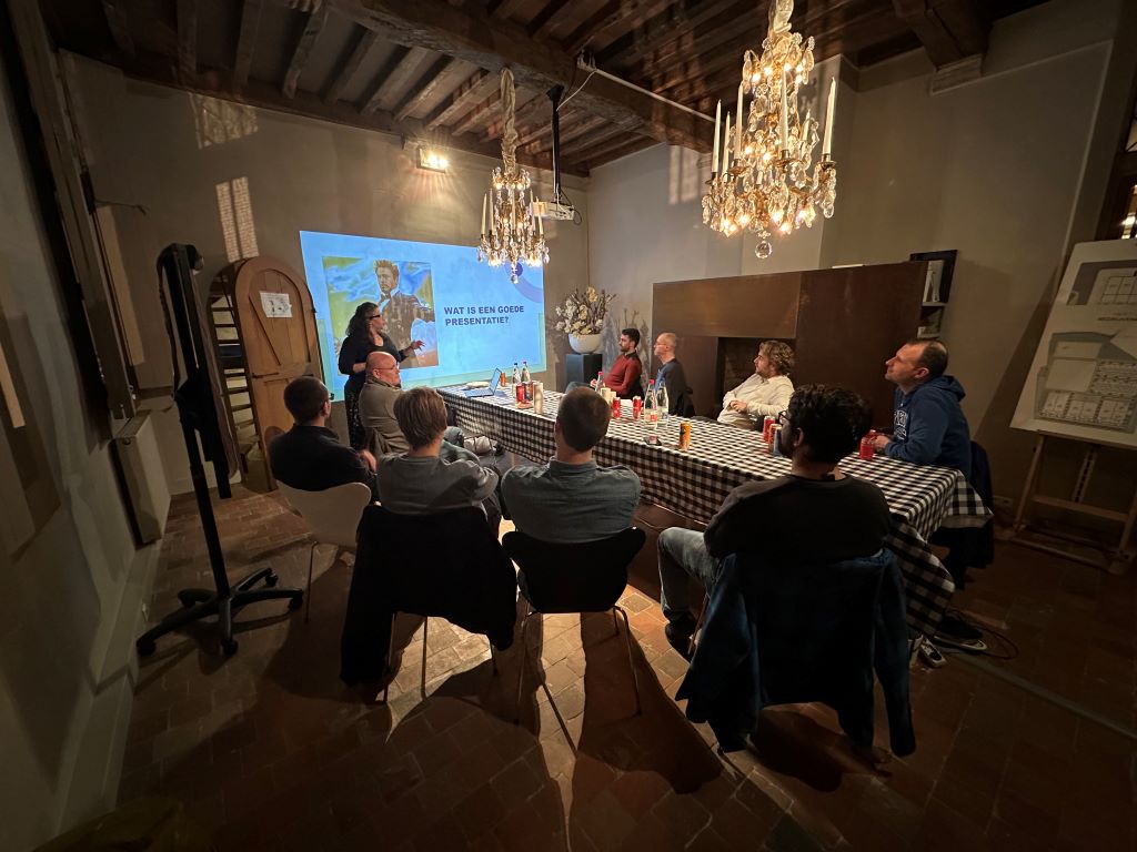 Participants are gathered around a checkered table in a cozy, rustic room with warm lighting and an elegant chandelier, setting the stage for a relaxed yet focused environment. Talisa leads the session at the front, presenting a slide titled *Wat is een goede presentatie?*, while attendees engage attentively, contributing to an interactive and collaborative learning atmosphere.